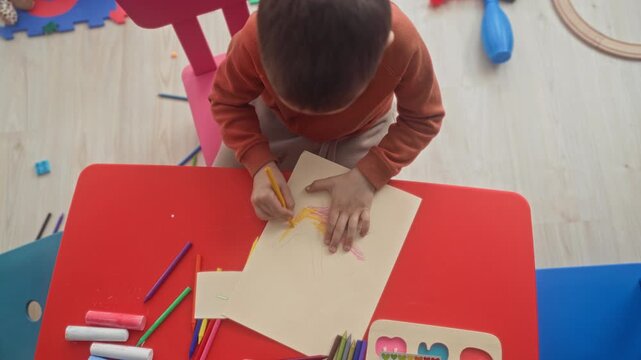 Boy holds yellow pencil and traces a hand outline on paper while seated at a red playroom table with scattered crayons and a pink chair visible; creativity concentration.