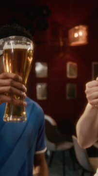 Vertical slowmo shot of joyful young multiethnic friends in team apparel raising beer glasses while celebrating sports win in bar together