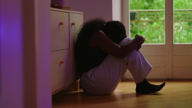 African American woman sitting on floor by window leaning forward with head down preoccupied with thoughts expressing worry concern and deep internal focus in quiet indoor space