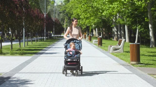 Sustained wide shot of a mother pushing her baby in a stroller in the park.