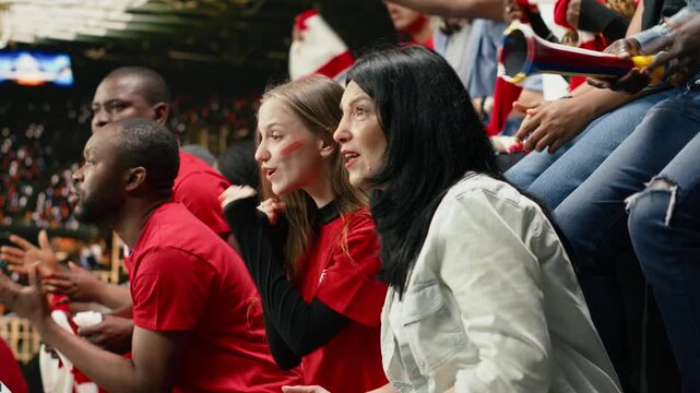 Excited female soccer fans leaning forward and cheering from stadium seats with diverse supporter crowd, red shirts and scarves during live match action, worried supporters focus on the game.