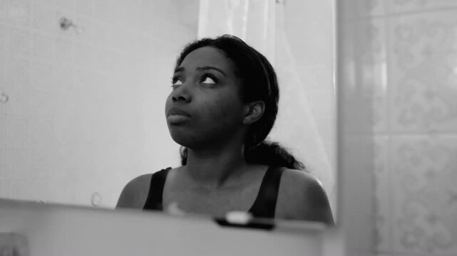 African American woman looking down at her reflection in bathroom mirror with sad and troubled expression creating emotional monochrome moment of self doubt and vulnerability in indoor setting