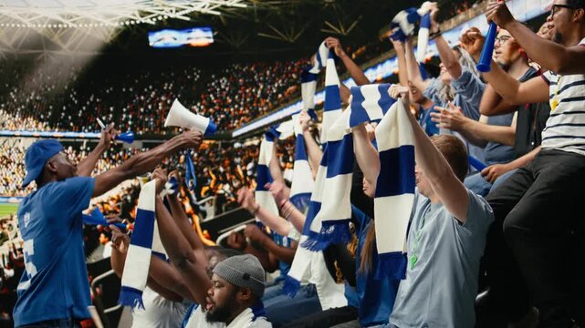 Excited blue team football supporters raising arms and cheering in packed stadium stands during thrilling live soccer match atmosphere, soccer fans shouting and singing chants, global event.
