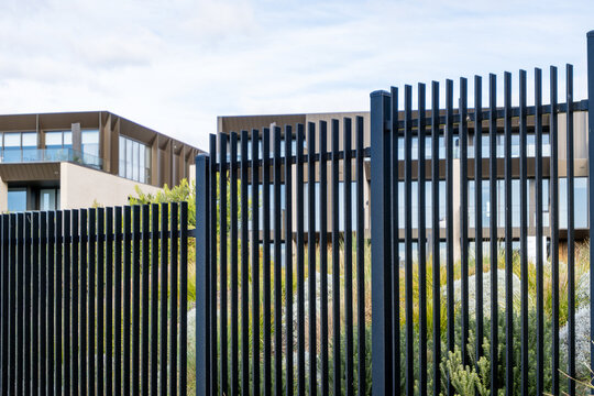 Close-up view of a sleek black steel palisade security fence with vertical slat design, set against a contemporary multi-storey residential home. Modern metal fencing in front of apartment buildings