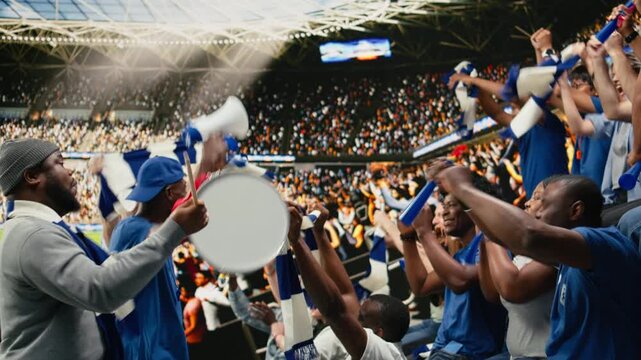 Dynamic football crowd leader motivating blue supporters with megaphone and drum during coordinated chant in packed stadium stands, organized supporter culture, diverse active fans.