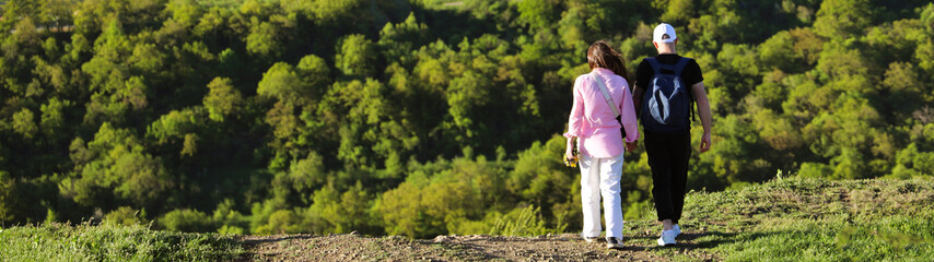 Obraz premium Rear back view of young couple hikers with backpacks rucksack holding hands and walking along a road with beautiful hills mountain with forest landscape with rural hone houses on background