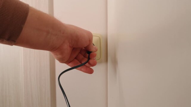 Close-up of hand plugging electrical cable into wall socket in modern residential apartment room.
