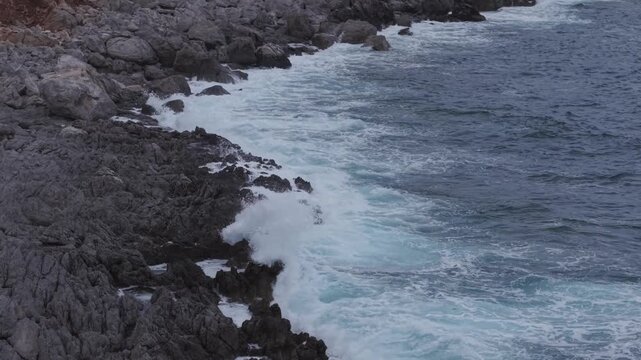 Strong ocean waves roll onto a rugged rocky shoreline, creating white sea foam and dramatic movement along the coast.
