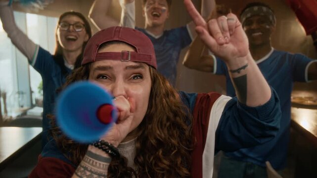 Slowmo portrait of young Caucasian girl in baseball cap looking at camera while blowing horn in sports bar, celebrating win with diverse friends cheering in blurred background
