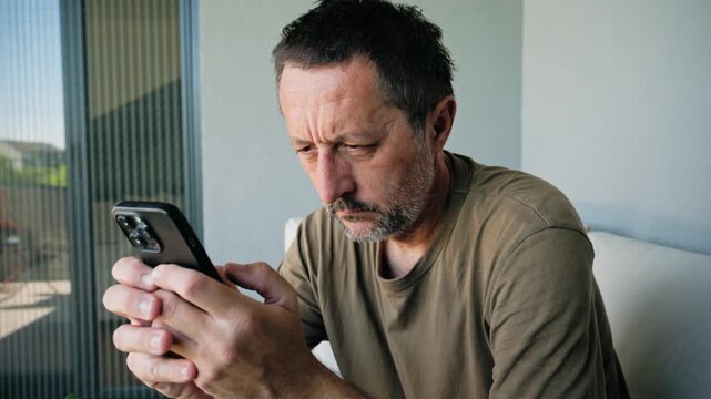 Middle-aged man sits on a balcony sofa near, intently looking at his smartphone with a worried expression, conveying focus, solitude and the everyday tension of modern digital life. 4K.
