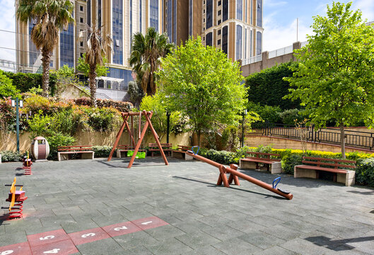 Children play area in urban park with swings and seesaw surrounded by trees and buildings during sunny day