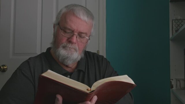 A man with a beard reads a book in a bright room. He sits quietly and focuses on the pages. Sunlight enters through the window. This takes place in the afternoon.