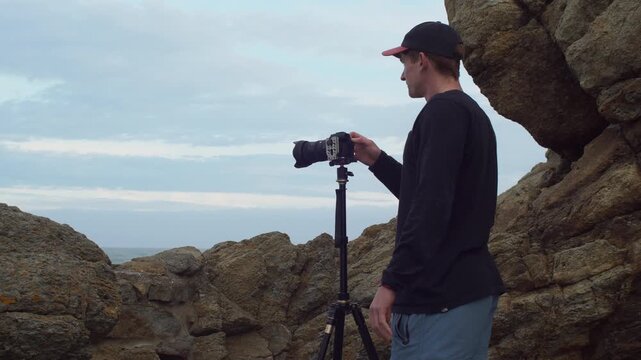 Man operating a professional video camera mounted on a tripod. Large rock formations in the foreground under a cloudy sky. Static medium eye level view.