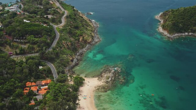 Thailand, Phuket coastline beach with winding road along lush green hills, turquoise sea, secluded sandy bay and orange roofed buildings. Summer travel tropical landscape. Aerial view drone flight