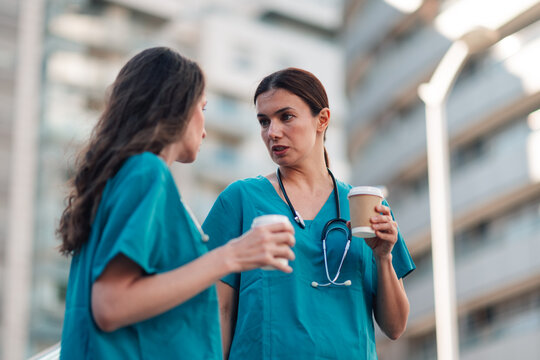 Two female nurses, wearing teal scrubs, having a coffee break outdoors, sharing thoughts and collaborating