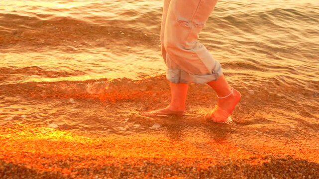 Beach walking sunset woman in denim pants strolls along the shoreline at golden hour as gentle waves lap at her bare feet