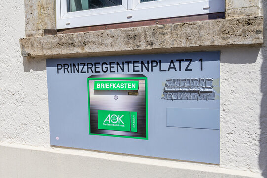 Augsburg, Germany - 25 May 2026: A close-up of a wall sign at Prinzregentenplatz 1 featuring an AOK Bayern health insurance mailbox under a window.