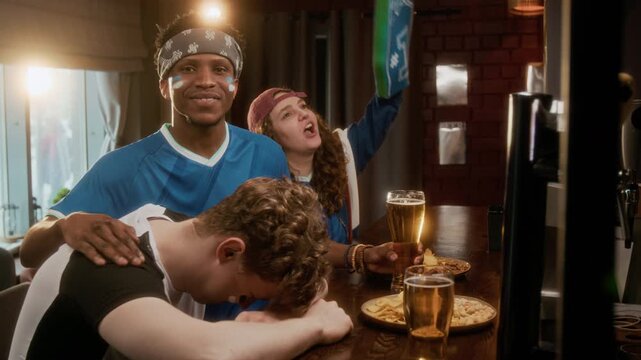 Handheld medium shot of young African American guy with painted face wearing blue team jersey consoling his male friend sitting at bar counter and feeling disappointed with sports match result