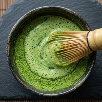 Overhead Close Up of Vibrant Green Matcha Powder Being Whisked into Frothy Tea in Dark Ceramic Bowl on Slate Coaster