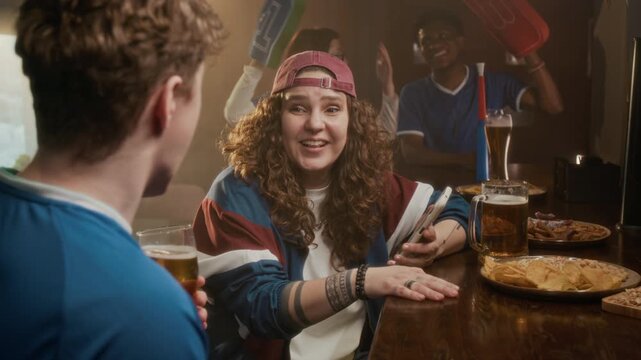 Handheld medium shot of excited young Caucasian girl in baseball cap sitting at bar counter and cheering while looking at sports betting app on smartphone, watching match with friends in pub