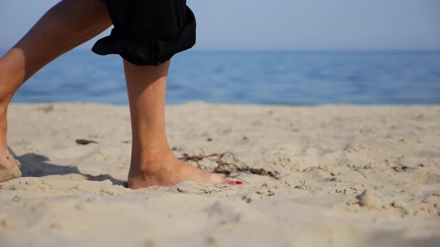 Female feet walking on sand at the beach with seascape at background. Legs of adult woman stepping along sea shore. Barefoot lady strolling near ocean coast. Concept of summer vacation. Low view