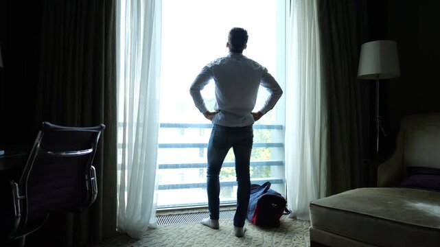 Tired young businessman in shirt standing near large window in modern hotel room or apartment interior after long trip. Exhausted manager resting after business travel showing fatigue and stress