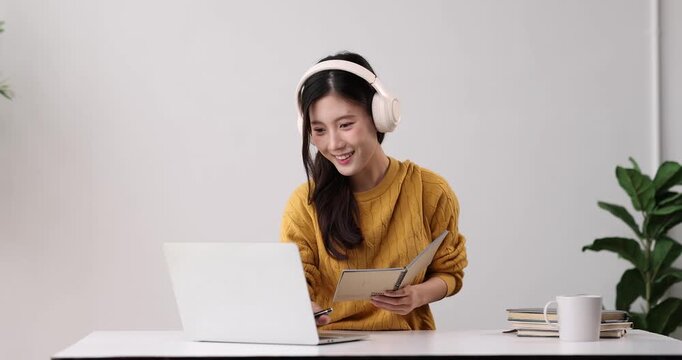 Young asian woman wearing headphones while diligently writing in a notebook, concentrating on her studies at home, engaged in self-learning and remote work with technology
