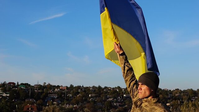 Ukrainian army man stands with raised national banner to peak of hill. Male soldier in military uniform with Ukraine flag as symbol of victory against russian aggression. Invasion resistance concept