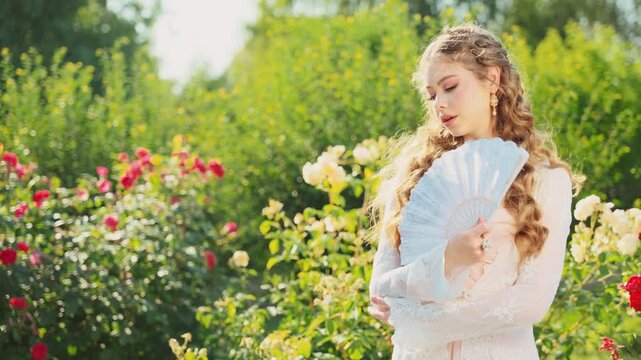 Portrait vintage lady girl medieval princess holding fan in hands waving enjoying coolness fresh air wind green summer nature garden roses. Young woman queen blonde old style white dress jewelry set