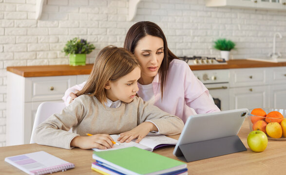 Portrait of a young child boy studying online class on tablet with his cheerful smiling mother at home. Woman help her son with homework during remote lesson. Distance education concept.