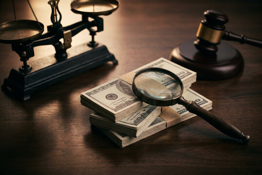 Magnifying glass resting on a stack of hundred dollar bills next to a wooden gavel and scales of justice on a desk