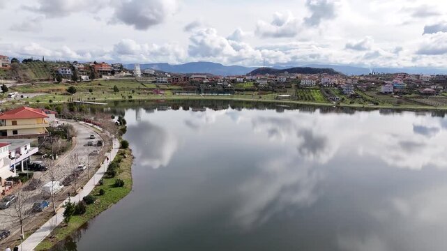 Wide aerial tracking shot flying along a paved lakeside pedestrian promenade, curved paths, and striking white cloud reflections on the glassy surface of Lake Belsh, Albania