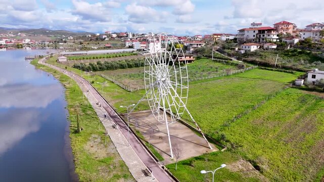 Aerial tracking shot circling a large white ferris wheel structure, revealing the scenic lakeside promenade, city skyline, and calm water reflections of Belsh Lake, Albania