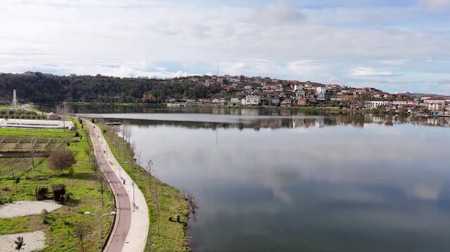 High-angle aerial panoramic establishing shot overlooking a winding pedestrian lakeside path, clear sky reflections on Lake Belsh, and a dense residential village on the opposite hillsides in Albania