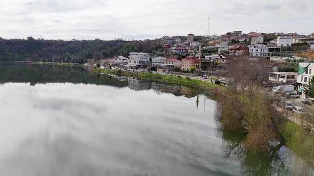 Wide aerial tracking shot flying along a lakeside road with parked cars, a modern wooden viewing deck platform with visitors, and peaceful village hillsides reflecting on Lake Belsh, Albania