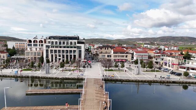 Aerial tracking shot moving forward over calm lake reflections toward a wooden pedestrian pier, a blue and white tour boat named Unique, and lakeside town buildings in Belsh, Albania