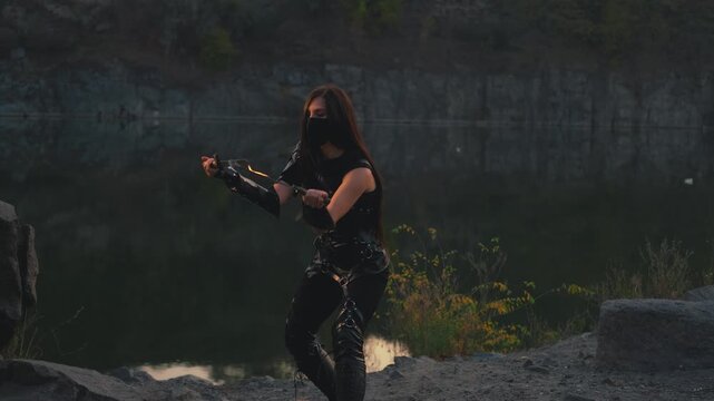  fantasy woman warrior assassin holds burning fire dagger in hands walks at night nature. Gothic sexy armed girl. Black leather costume mask hides face. Ninja sword knife. 4k footage. river rocks