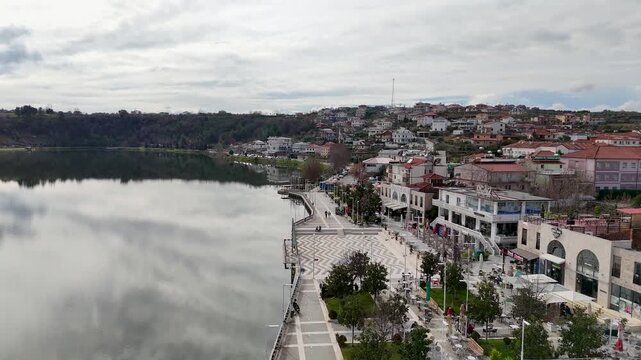 Aerial tracking shot moving forward over the glassy surface of Lake Belsh, past a patterned pedestrian promenade plaza, outdoor cafe umbrellas, and hillside houses in Albania