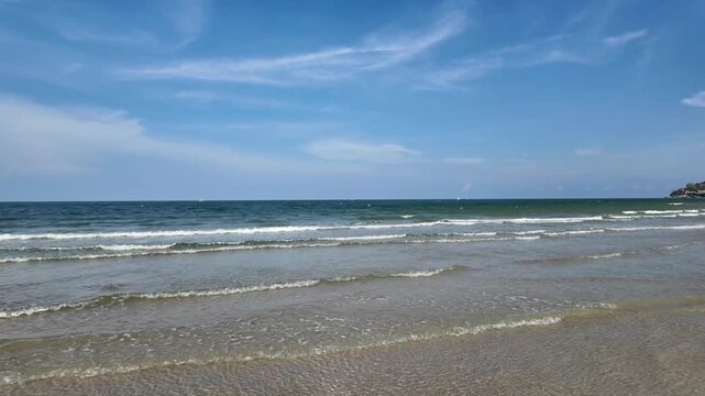 Serene Ocean Vista: Gentle waves lap at a sandy shore under a vast blue sky dotted with wispy clouds, offering a tranquil seascape.
