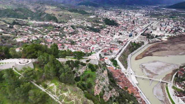 Wide aerial tracking shot flying forward over the steep green cliffs and stone bastions of Berat Castle, revealing the Osum River, modern city grid, and distant mountain ridges in Albania