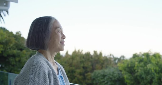 Senior Asian woman sitting balcony rail soft light causing head turning and smiling, copy space