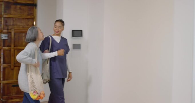 Caregiver opening wooden door and escorting senior with mesh bag toward kitchen, showing layout