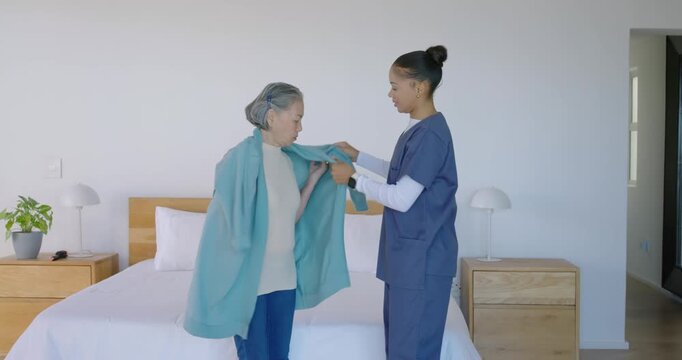 Senior Asian woman and female aide lifting jacket guiding sleeves adjusting for warmth in bedroom