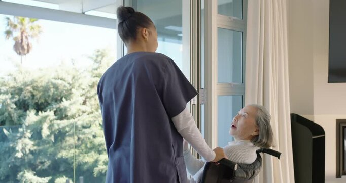 African American aide steadying wheelchair, guiding senior Asian woman to glass door to view garden