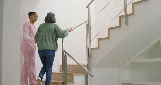 Carer in pink scrubs taking Senior woman's arm, guiding up wooden stairs using handrail for balance