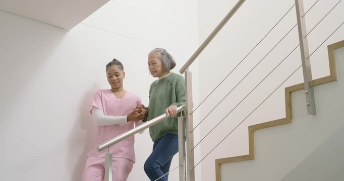 Female caregiver in pink scrubs guiding senior reaching handrail, descending stairs for support