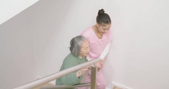 Woman aide in pink scrubs offering hand guiding Asian senior down stairwell steadying on metal rail