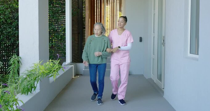 African American nurse in scrubs escorting senior Asian woman reaching for planter on covered porch