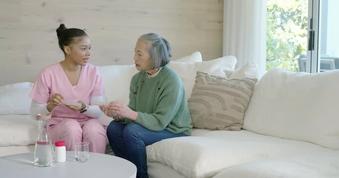 Female caregiver presenting blister pack, showing dosage to senior Asian woman on couch, copy space