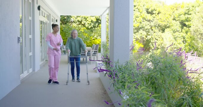 Senior female using walker and aide in pink scrubs walking porch pots drawing senior leaning blooms